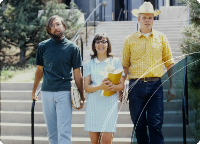 Three Central Oregon Community College students from the 1960s era walking down steps outside. The student on the left is wearing a green turtleneck and light-colored pants, the middle student is wearing glasses and a light blue checkered dress, and the student on the right is wearing a yellow plaid shirt, blue jeans, and a cowboy hat. They are all carrying books and folders.