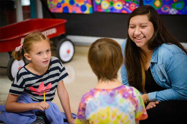 A woman kneels on the floor talking with two young children in a colorful classroom