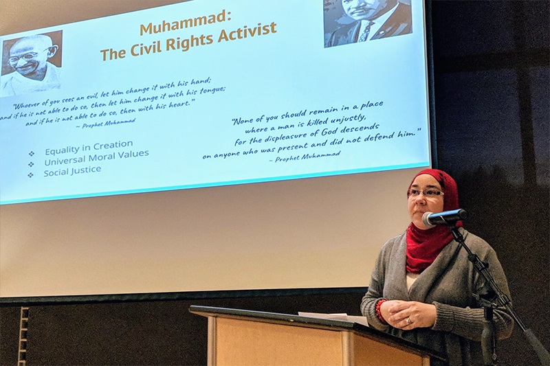 a woman wearing a hijab stands at a podium in front of a slide projecting the images of Ghandi and MLK Jr.