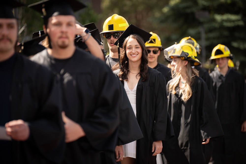 students in caps and gowns standing with some wearing yellow firefighting helmets 