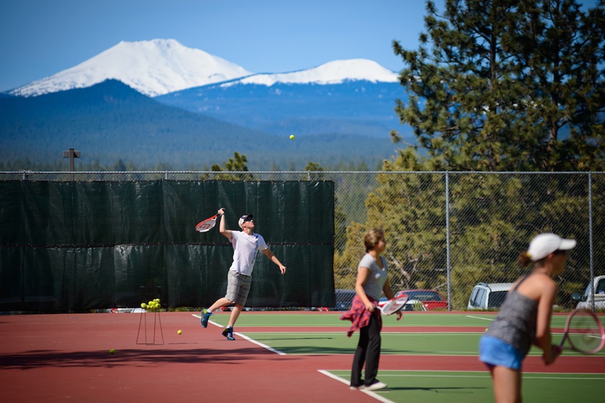 tennis player serving with red and green court and snowy mountains in distance