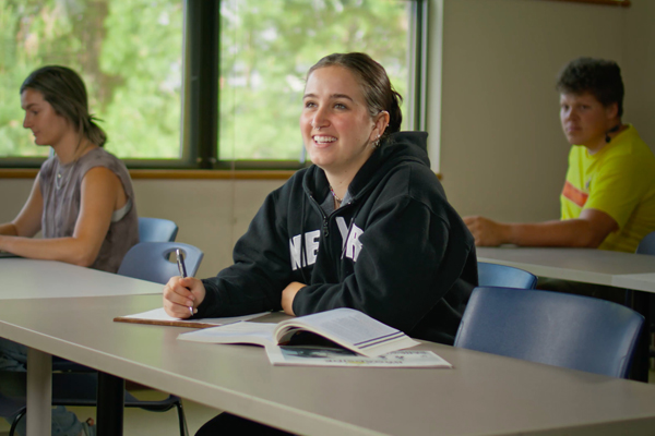 Central Oregon Community College Humanities students learning in classroom. One female student smiling in the middle with a female and male student in the background.
