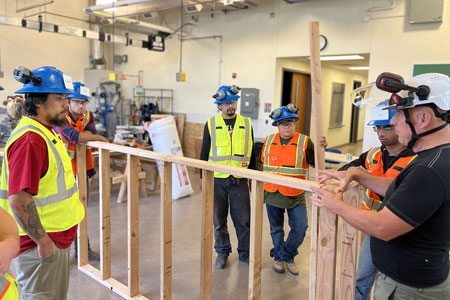 Hands on apprentice learning environment with teacher and students gathered around wooden construction framing.