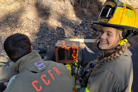 Female and male fire student studying a burn pattern lab. Femal student is looking towards camera and smiling with yellow fire helmet on.